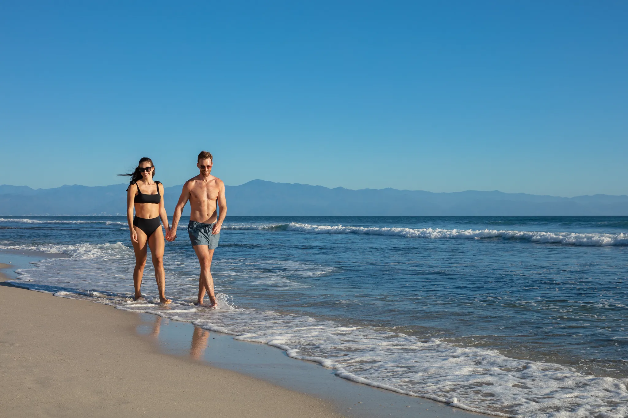 Couple holding hands while walking along the shoreline at Secrets Bahia Mita Surf & Spa Resort with ocean waves in the background.