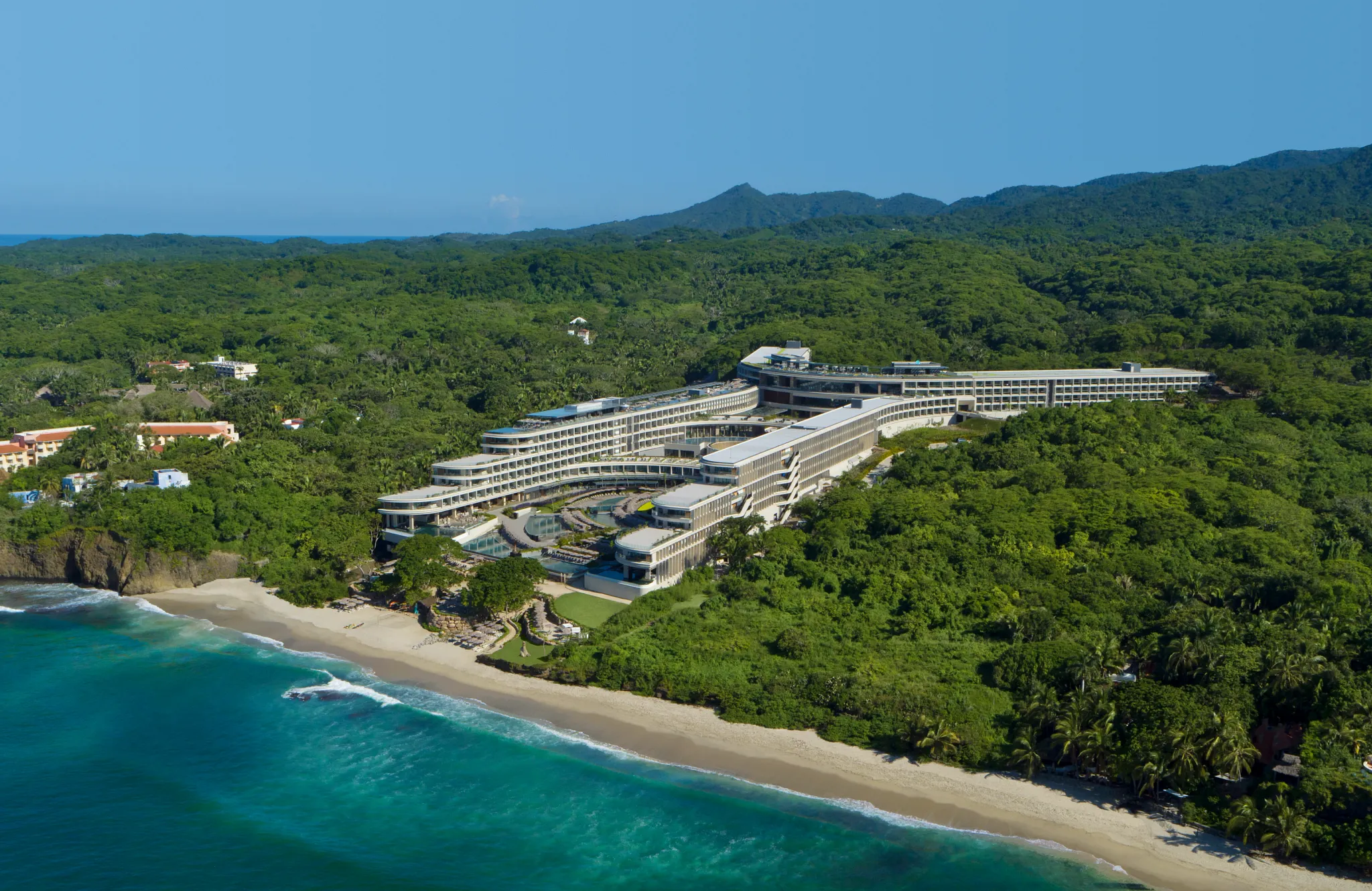 Panoramic aerial view of Secrets Bahia Mita Surf & Spa Resort along a secluded beach surrounded by lush greenery.